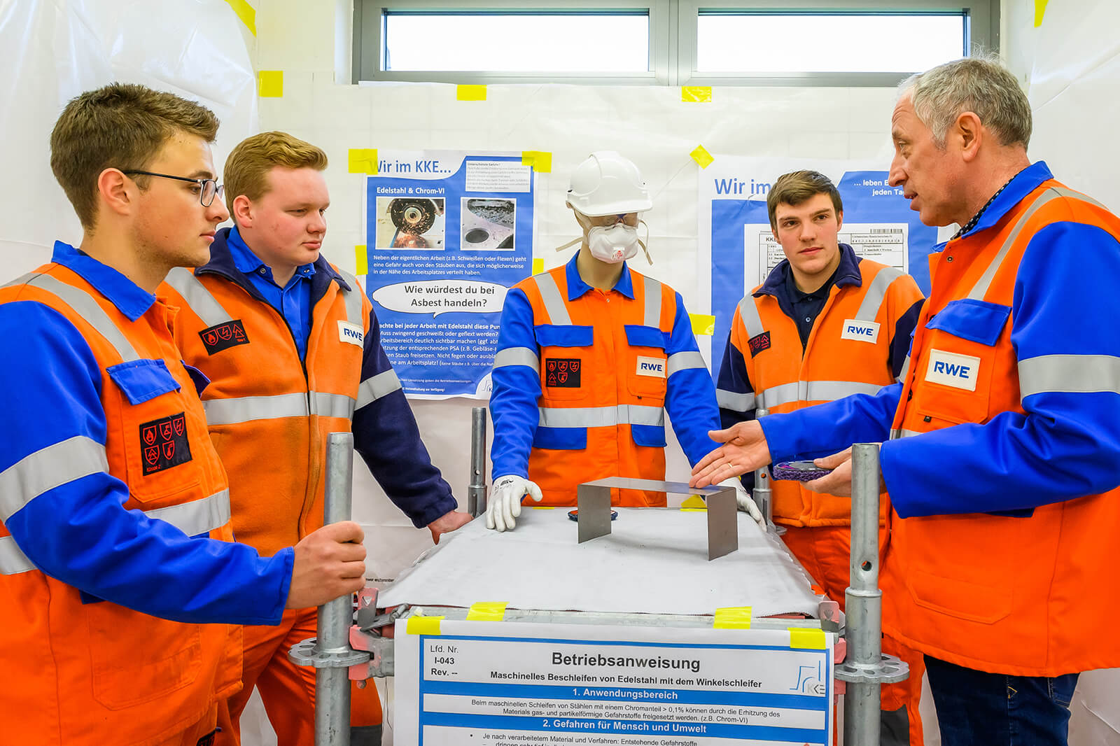 Fünf Personen in orangefarbenen Sicherheitswesten diskutieren über einen metallischen Gegenstand auf einem Tisch.