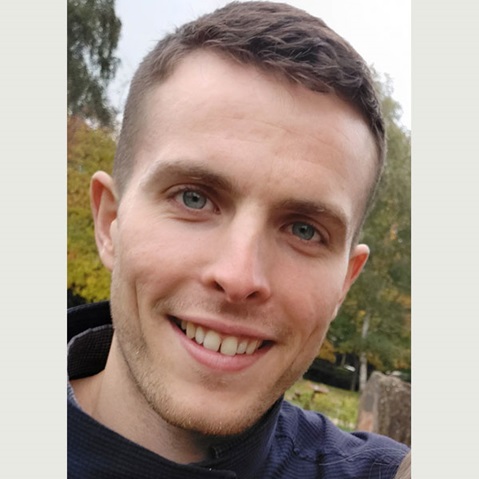 Eine Person mit einem verschwommenen Gesicht trägt ein dunkles Oberteil und steht im Freien zwischen Bäumen mit Herbstlaub.