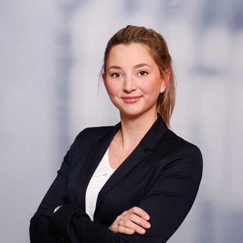 Eine Frau in einem schwarzen Blazer steht mit verschränkten Armen vor einem unscharfen Hintergrund.